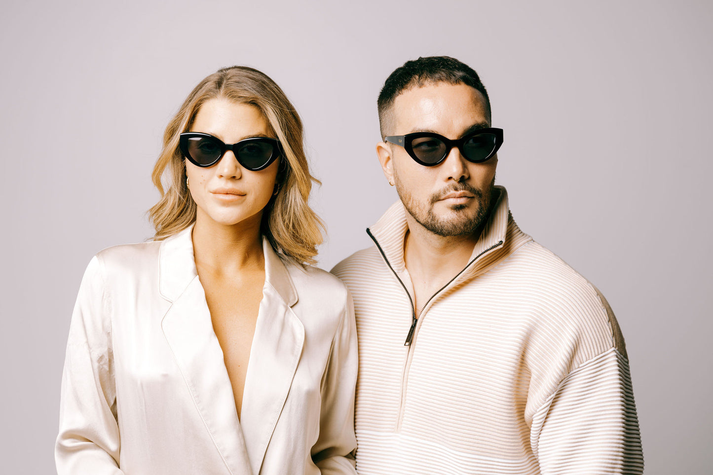 Woman and man in cream clothing both wear Cleo charcoal black biodegradable sustainable cateye sunglasses with dark grey lens