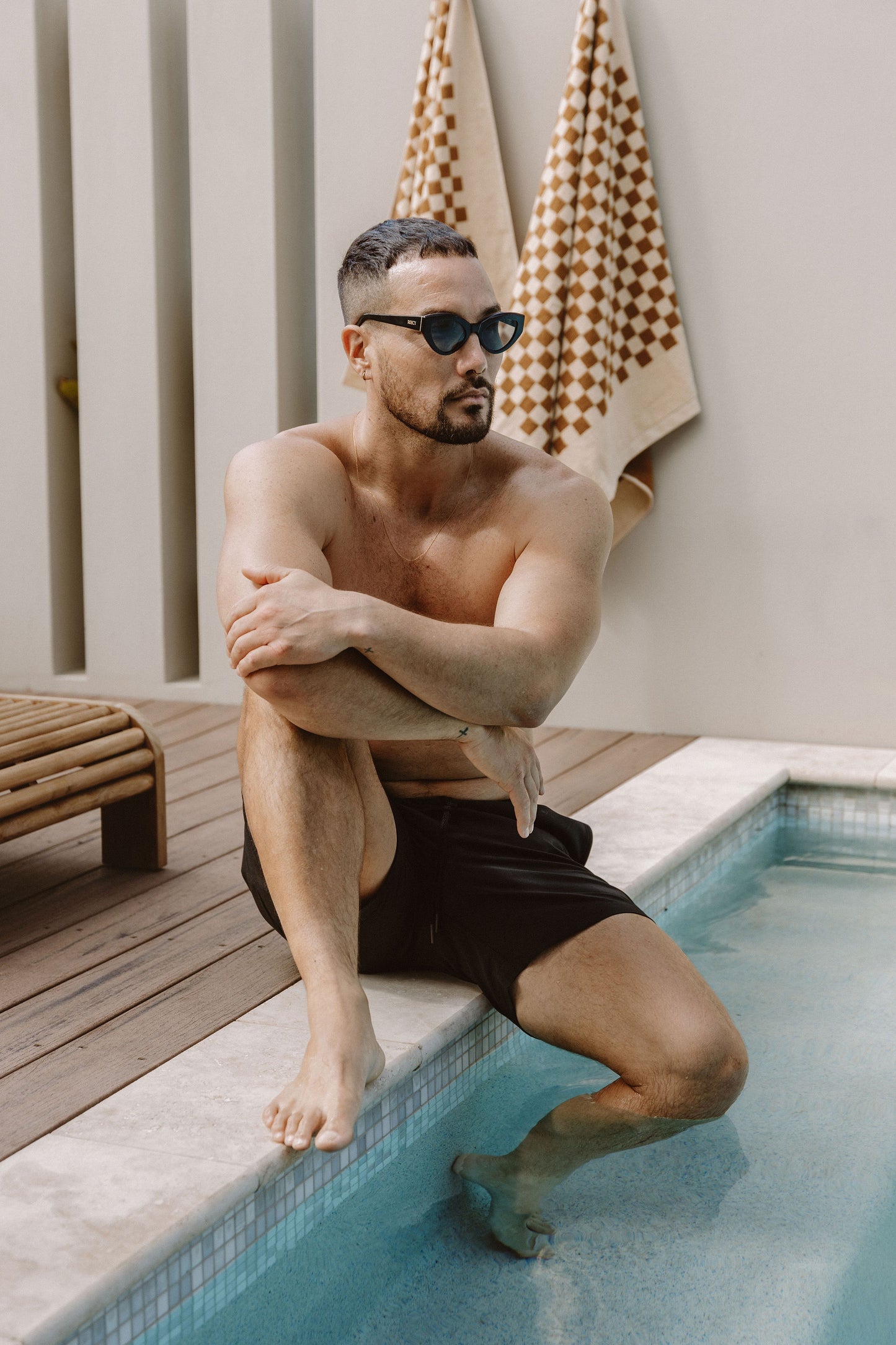 Man sitting by swimming pool in black shorts wearing Cleo charcoal black biodegradable sustainable cateye sunglasses with dark grey lens