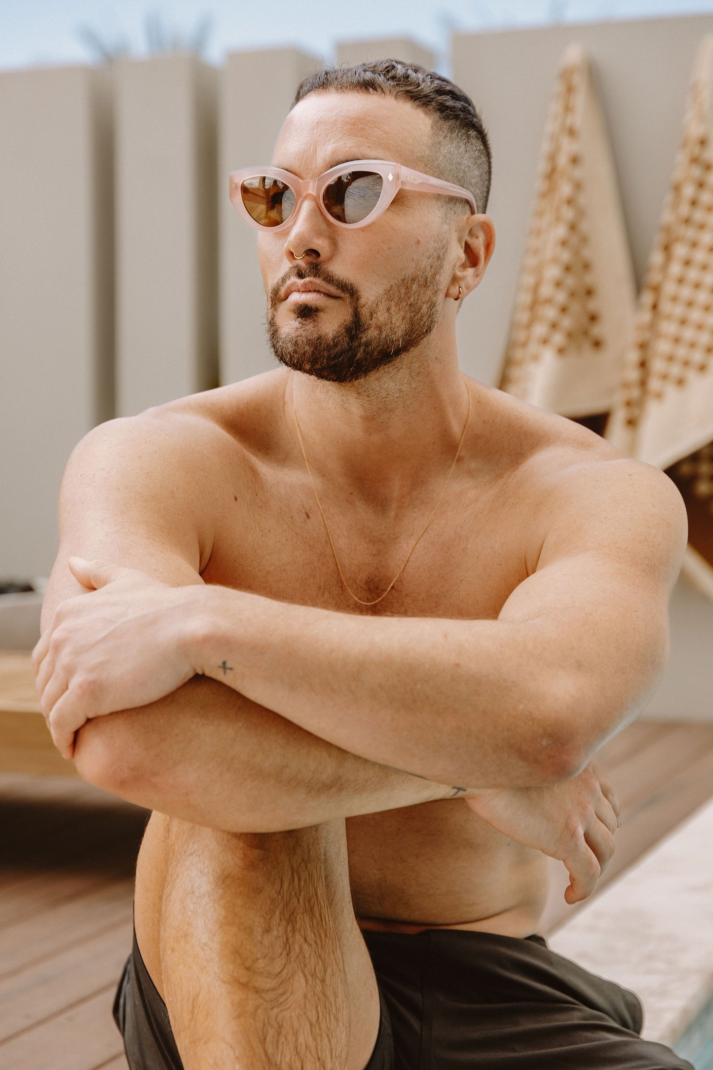 Man in black shorts sits by a swimming pool wearing pink Cleo Floss cateye biodegradable and sustainable sunglasses with tan lens