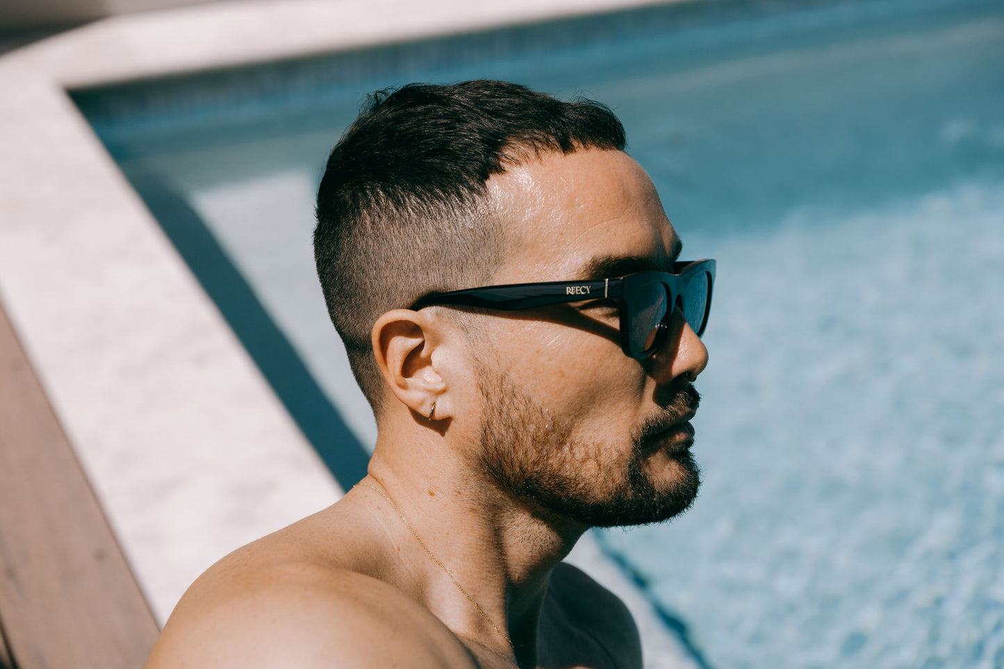 Man by a swimming pool wears black Ike Charcoal classic biodegradable and sustainable sunglasses with dark grey lens