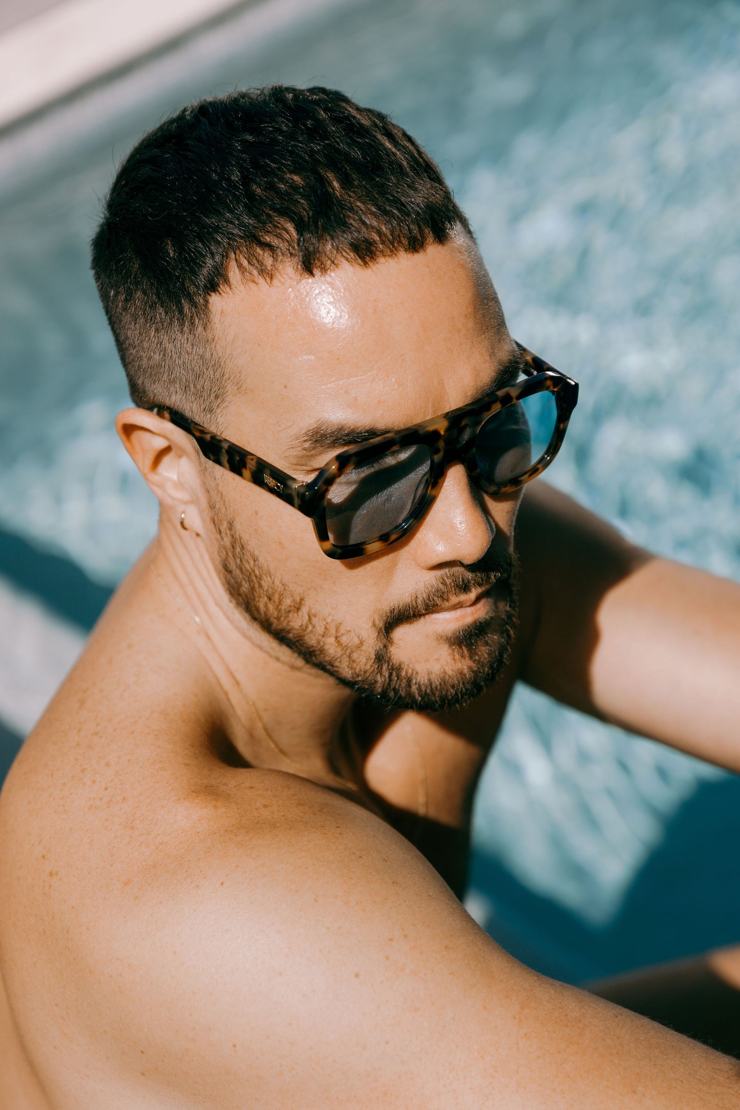 man without shirt wears dark tortoiseshell aviator biodegradable and sustainable malone brulee blue sunglasses with blue lens by a swimming pool